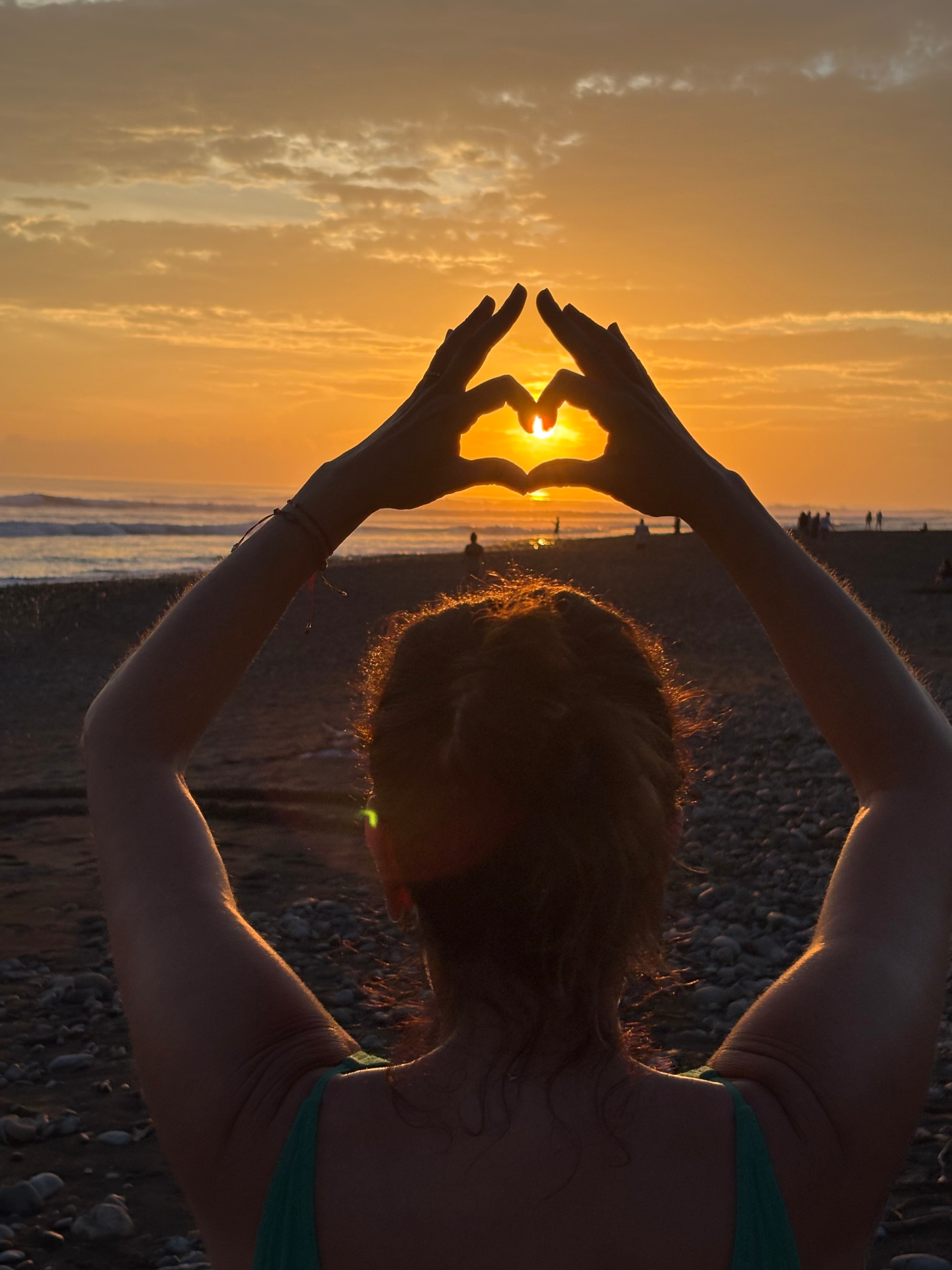Two hands forming a heart shape around the setting sun on a quiet beach